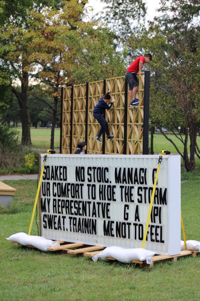 Image: Titus Wonsey's sculpture Is This It can be seen in the foreground. It is a marquee sign that says "SOAKED NO STOIC. MAAGI G UR COMFORT TO HIDE THE SOTMR MY REPRESENTATIVE GA A IPI SWEAT, TRAININ ME NOT TO FEEL." Kids climb on a wooden structure in the background. Photo by Carlos Flores. 